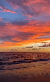The image captures a stunning beach scene at sunset. The sky is filled with dramatic, vibrant colors—deep oranges, pinks, and purples blend seamlessly with patches of blue, creating a breathtaking display. Wispy clouds are illuminated by the setting sun, casting warm hues across the horizon.